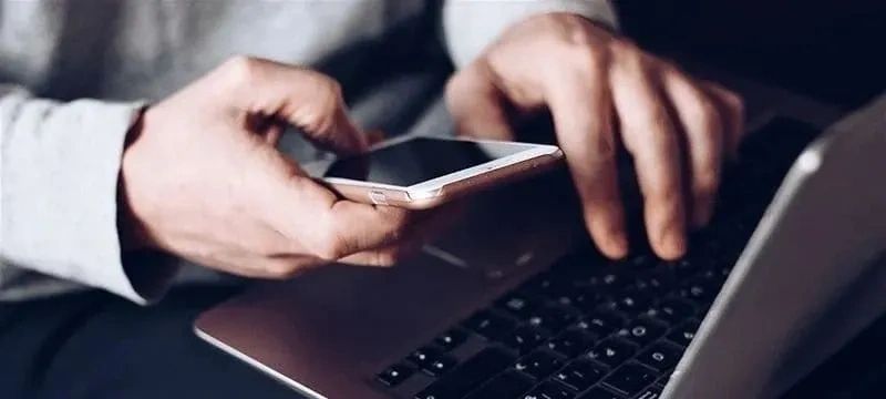 Person using a smartphone and typing on a laptop simultaneously.