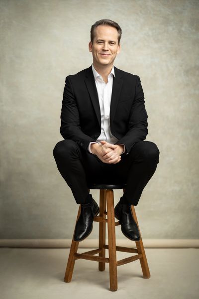 Man in a black suit sitting on a wooden stool.
