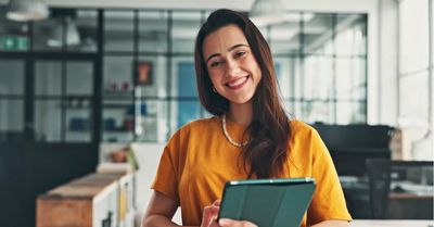 A woman in an open space office, smiling, holding a tablet device.