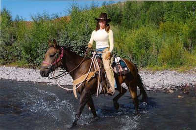 Woman in cowboy attire rides horse through shallow river.