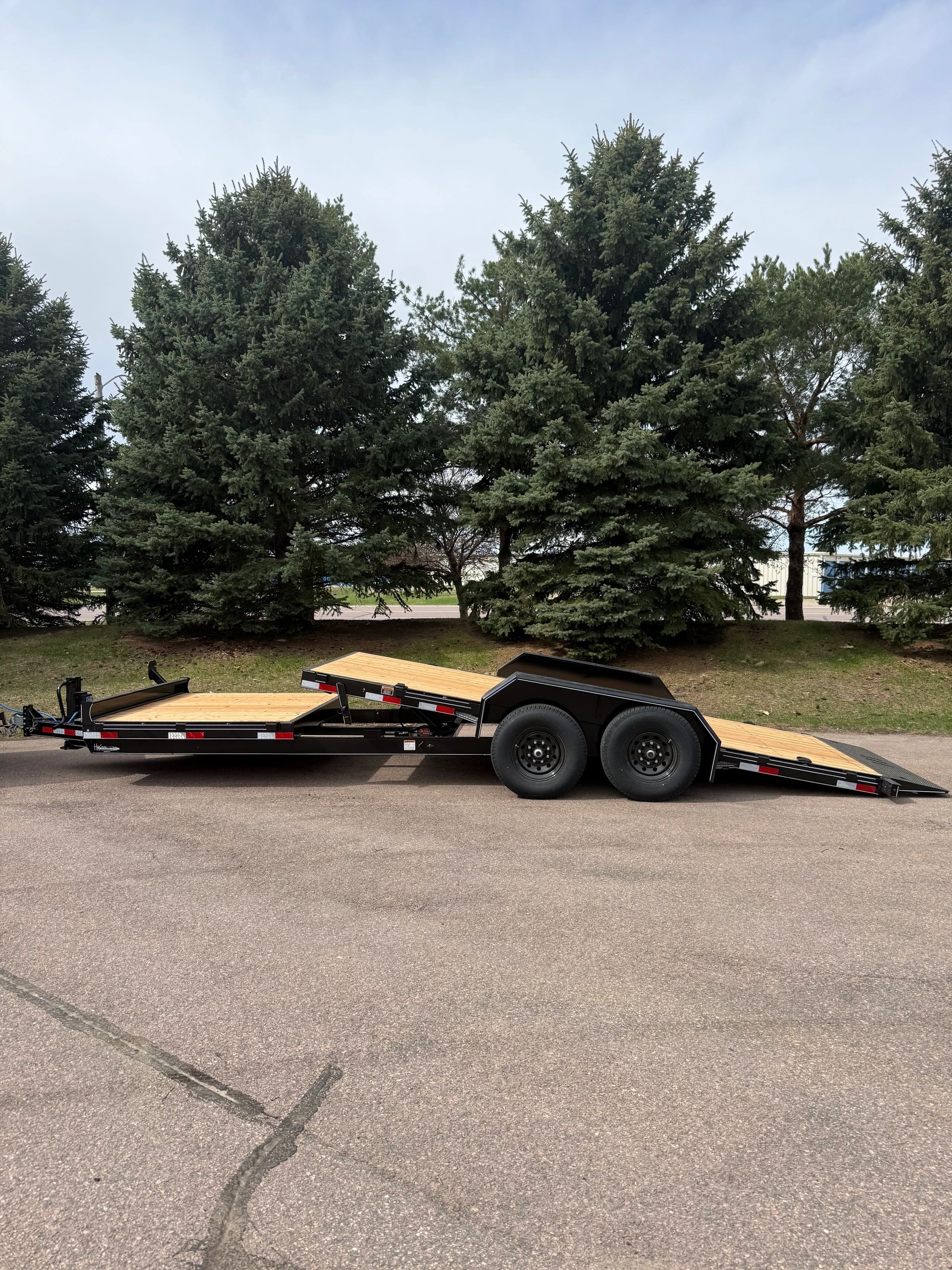 Black flatbed trailer with wooden deck parked on pavement near pine trees.