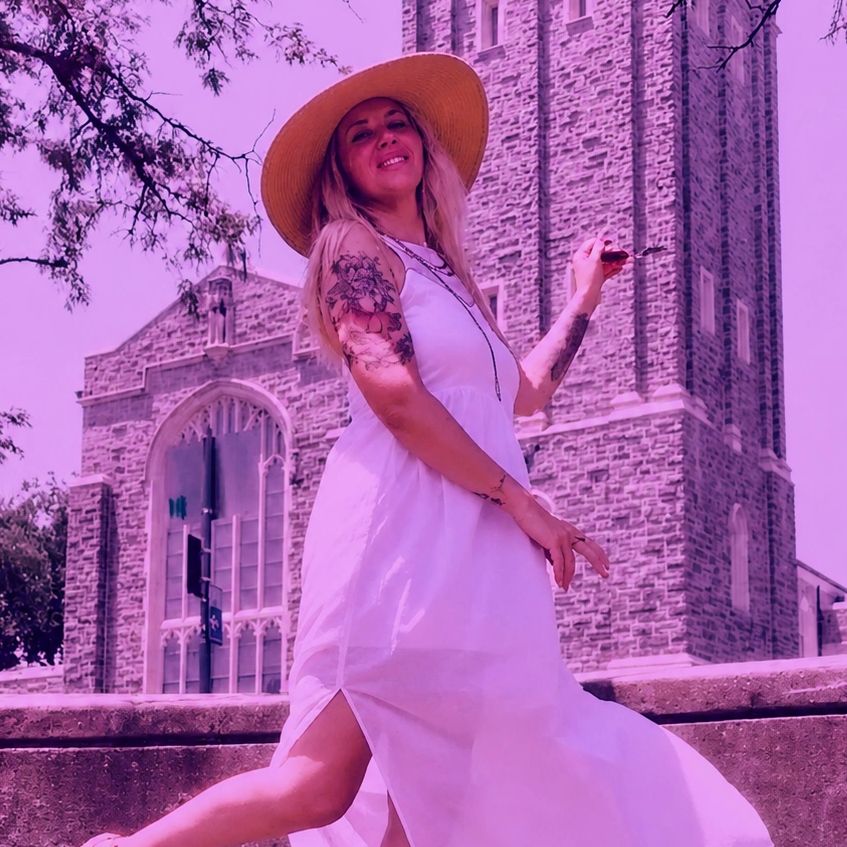 Woman in white dress and hat smiling in front of a stone building.