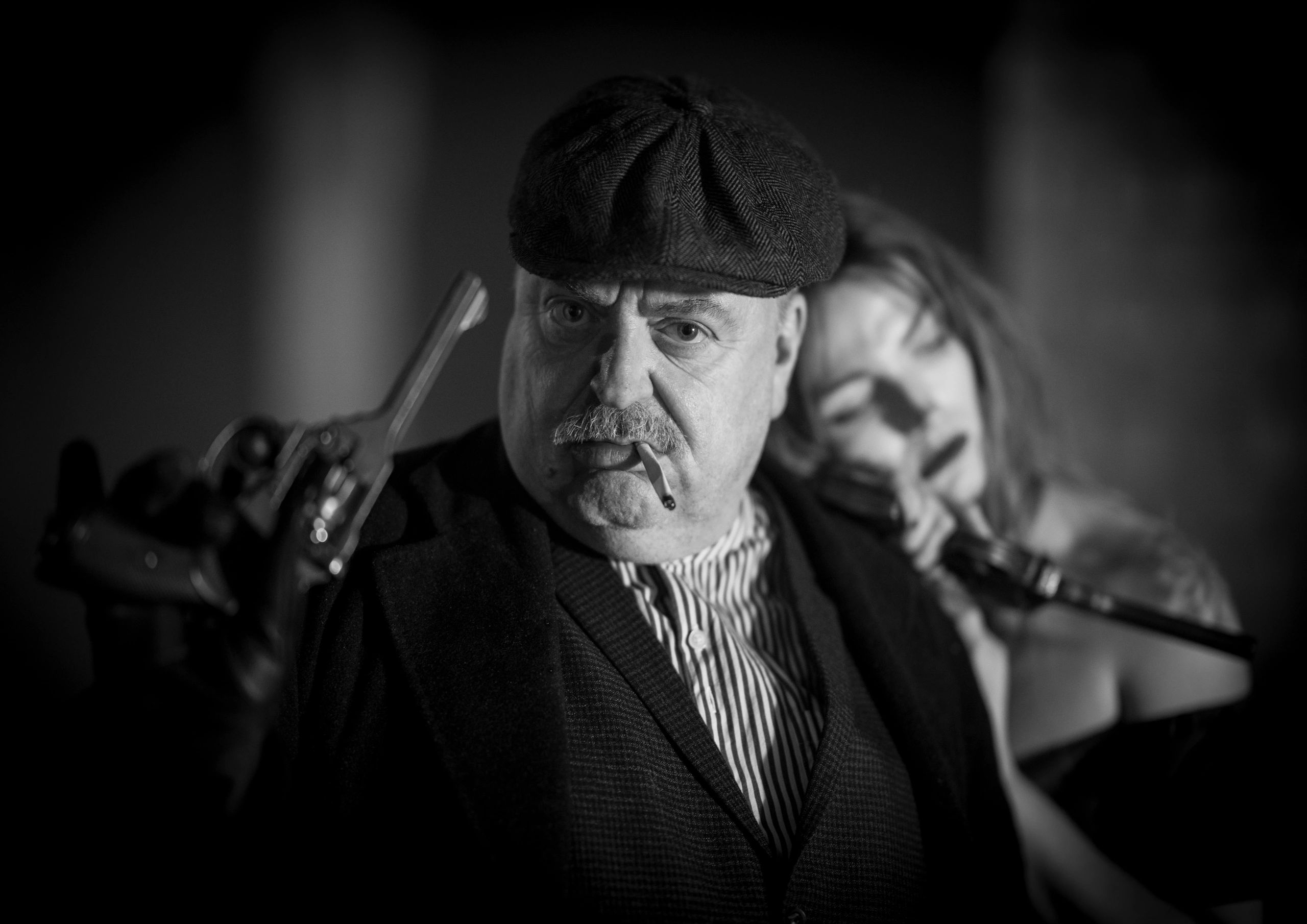 Man with a cigarette aiming a gun, woman behind him blurred.
