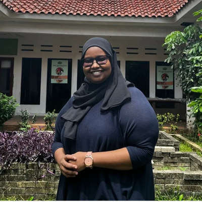 Smiling woman in a black hijab and glasses stands outdoors in front of a building.
