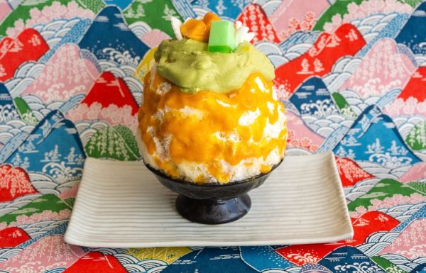 Colorful shaved ice dessert with mango and green toppings on a patterned background.