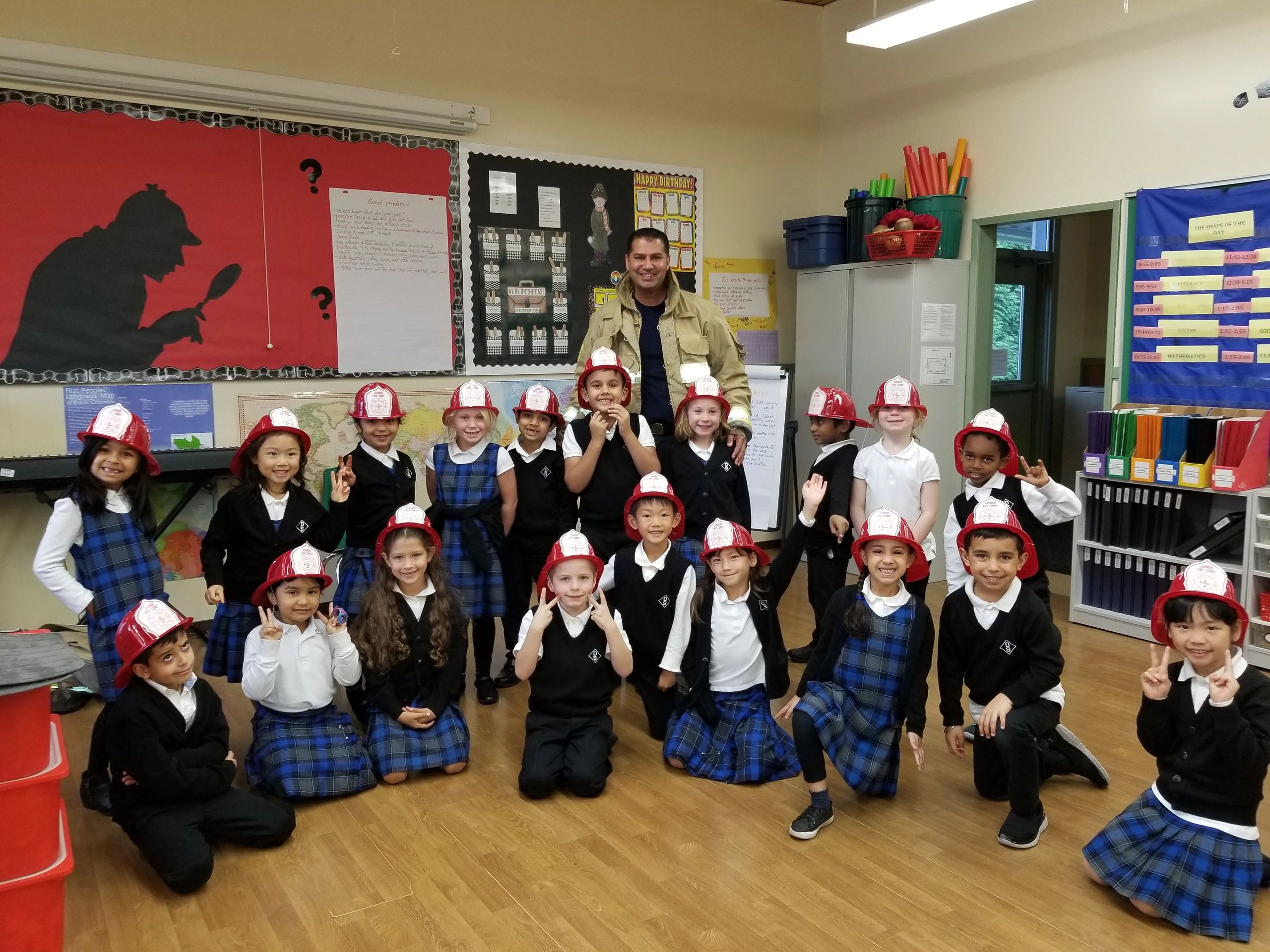 A young class wearing school uniforms and plastic firefighter hats pose with a visiting firefighter