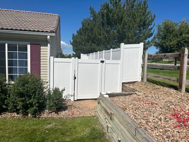 White vinyl fence gate next to a house with landscaping and blue sky.