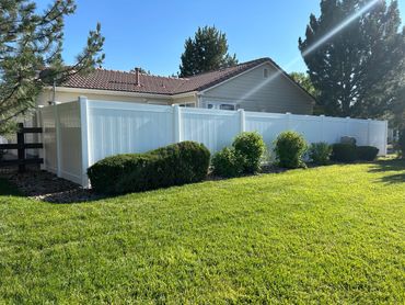 A suburban house with a white vinyl fence and green shrubs in a sunny yard.