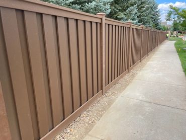 Long brown fence running alongside a sidewalk with trees in the background.