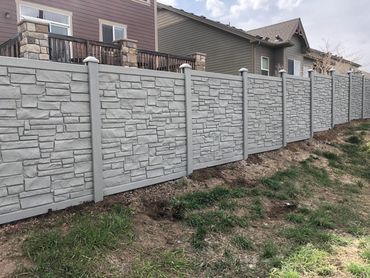 A long, gray stone-patterned fence separates two residential backyards.