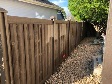 A brown wooden fence along a rock-covered yard with a tree and house in the background.