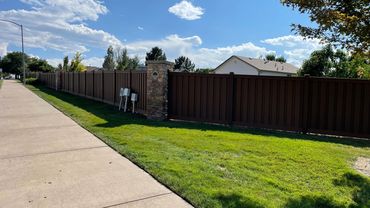 Long brown fence with stone pillars along a sunny sidewalk and green grass.