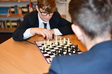 A student in a school blazer and glasses focuses intently while making a move during a chess match.