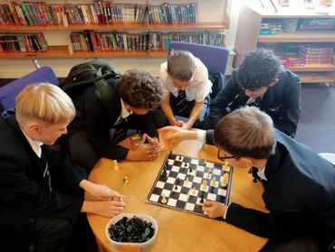 Five students in school blazers and ties gathered around a circular wooden table playing chess.