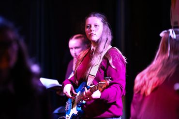 Student performer in a school uniform plays a blue bass guitar under purple stage lighting
