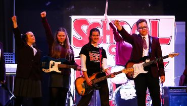 Student performers with guitars raise their fists in front of a "School of Rock" stage banner