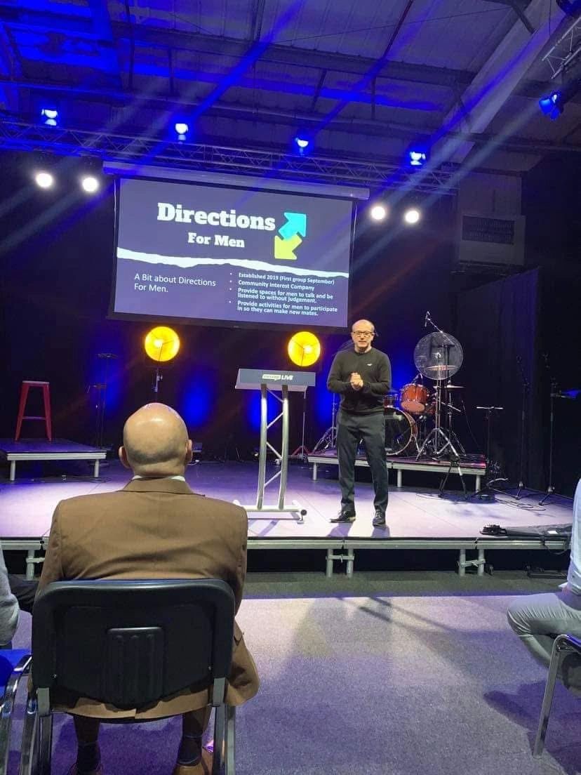 Man speaking on stage at a Directions For Men event with audience seated.