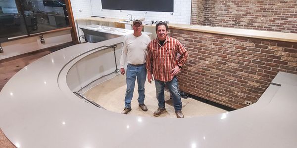 Two men stand inside a large, newly installed curved countertop in a modern kitchen or bar area.