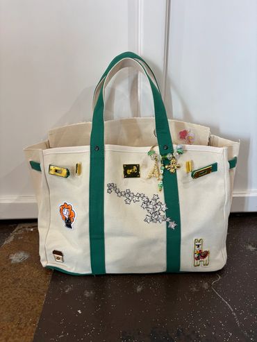 Beige tote bag with green straps, decorated with patches and charms.