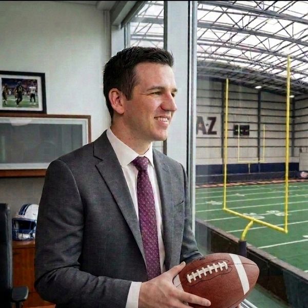 Man in suit holding a football by an indoor field window.