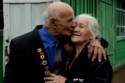 Very old couple with husband embracing and kissing wife and wife smiling with hand on his chest