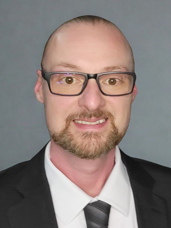 Smiling man with glasses in formal suit and tie against gray background.