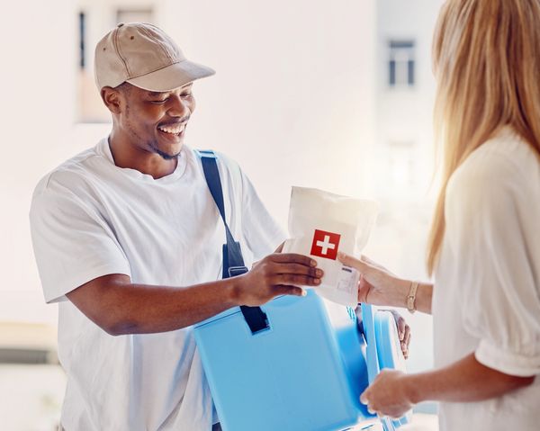 A person in a cap and white t-shirt hands a package with a red cross to another person.