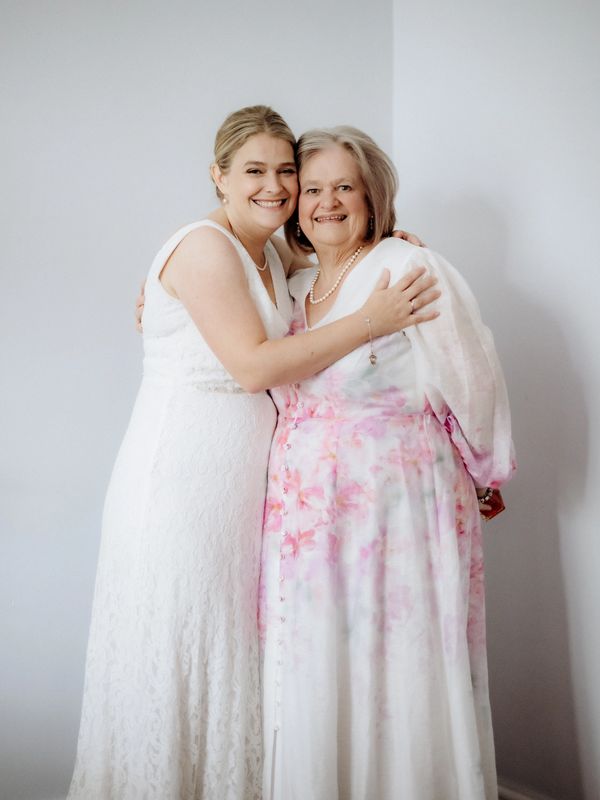 A bride and an older woman in floral dress smiling and hugging indoors.