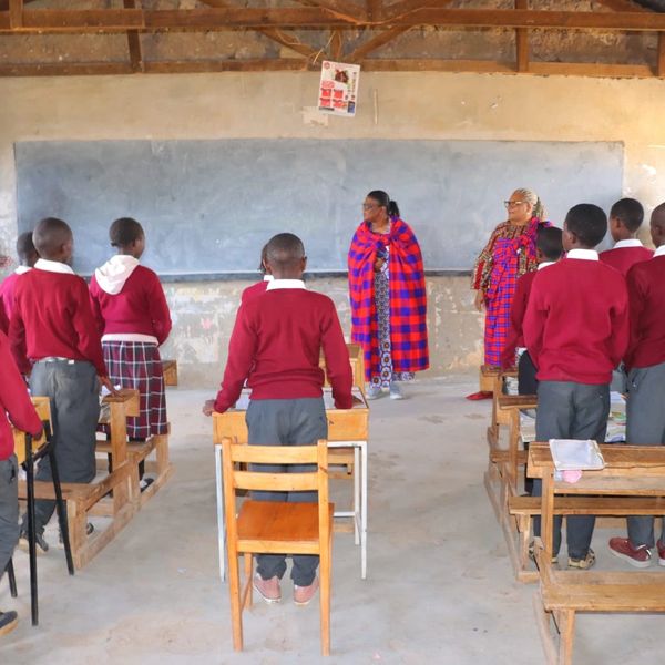 Students in red uniforms stand in a classroom with two women in traditional Maasai attire.