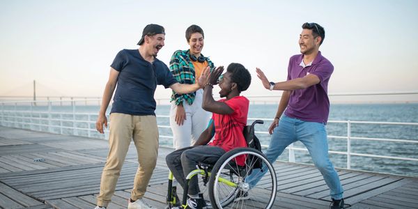 Groupe de jeunes gens s'amusent en entourant un jeune homme noir assis dans un fauteuil roulant.