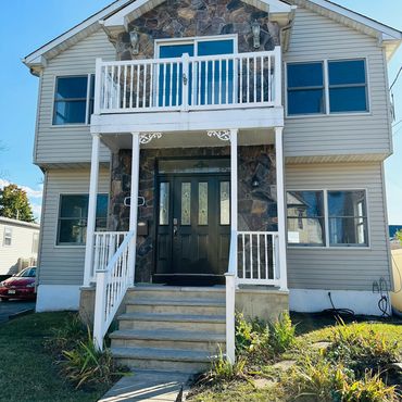 Two-story house with stone facade, black front door, and white balcony railing under clear blue sky.