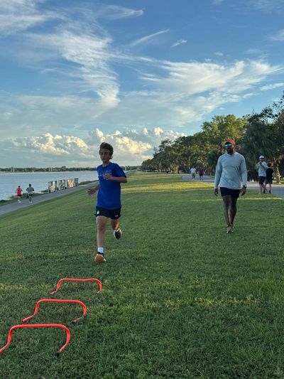 A boy runs between hurdles on a grassy path near water.