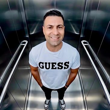 Man smiling in a metallic elevator wearing a white Guess t-shirt.