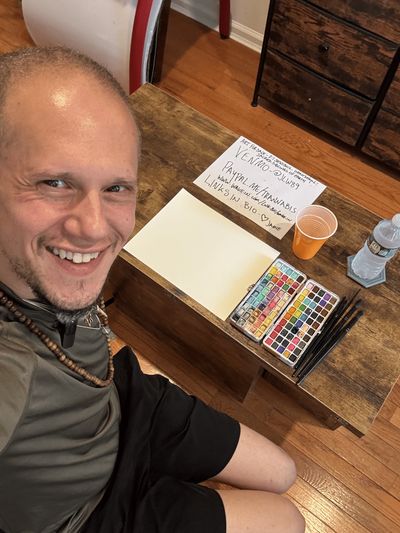 Jamie smiling looking up green shirt/black shorts, seated at table with watercolors set-up