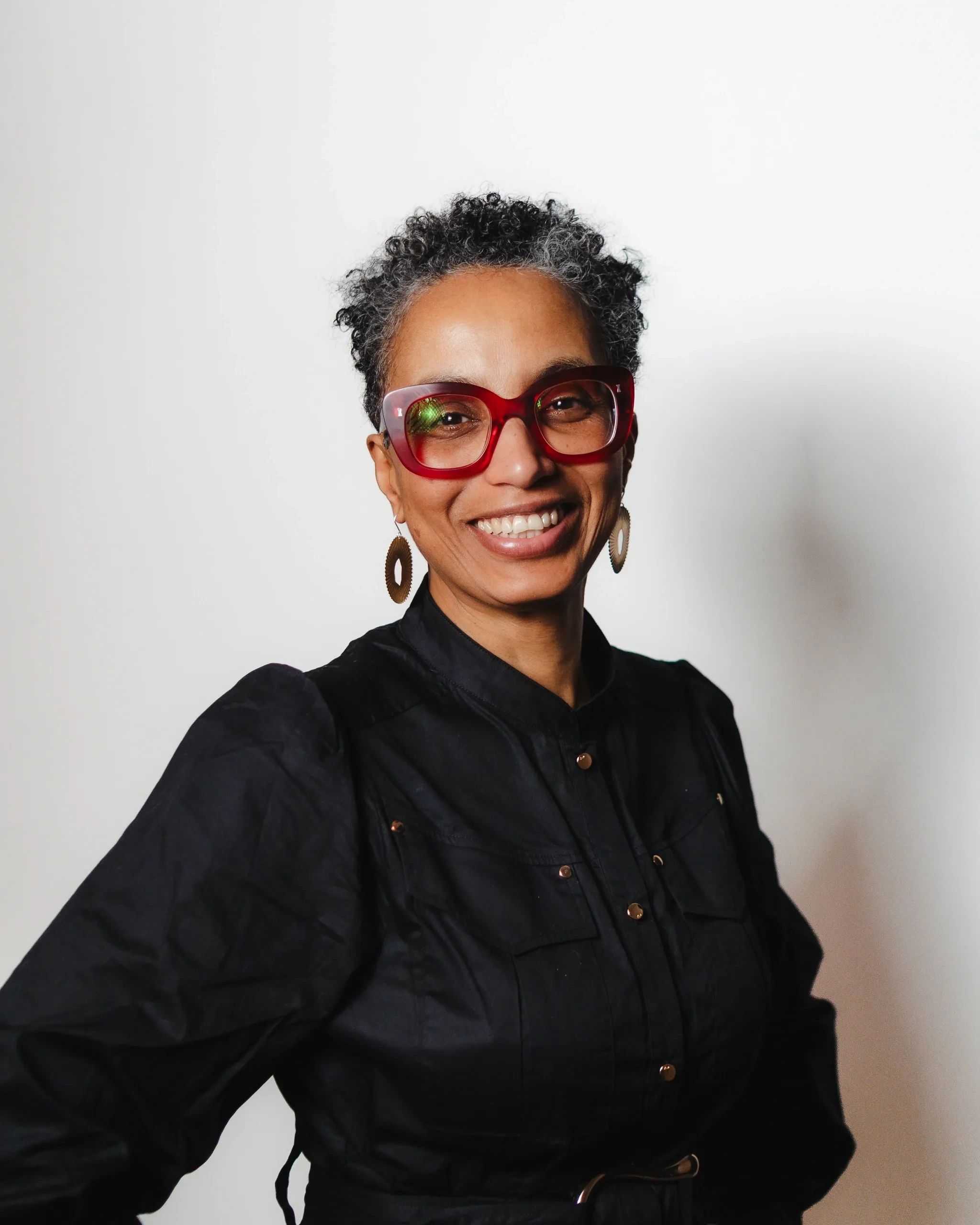 Mixed race woman in her early fifties wearing red coloured glasses, with golden earrings, short cut
