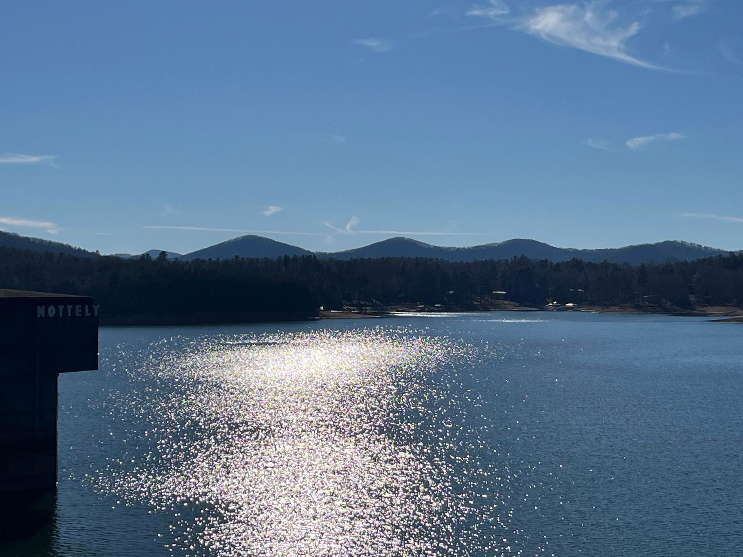 Sunlight sparkles on a calm lake with mountains in the background under a clear blue sky.