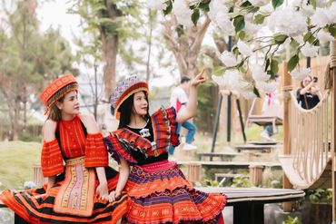 Two women in colorful traditional attire admire white blossoms outdoors.