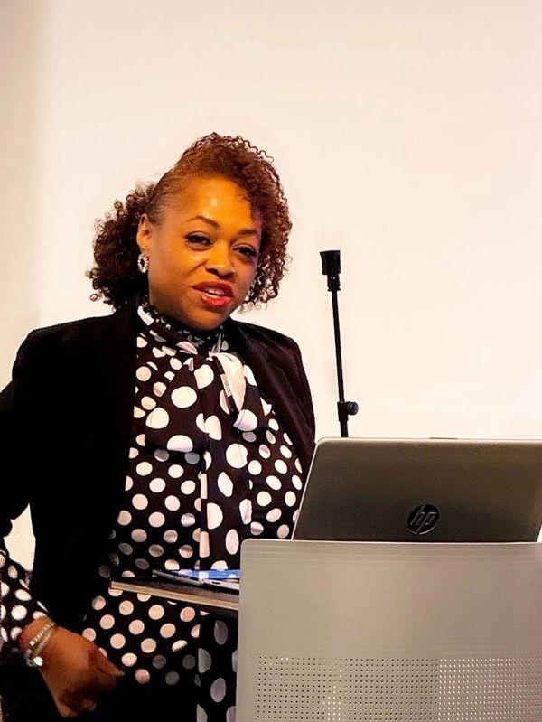 Woman in polka dot blouse speaking at a podium with a laptop.
