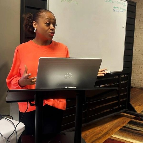 Woman in orange blouse giving a presentation with a laptop and whiteboard.