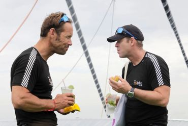 Two men in black shirts enjoying drinks and fruit on a catamaran trip.