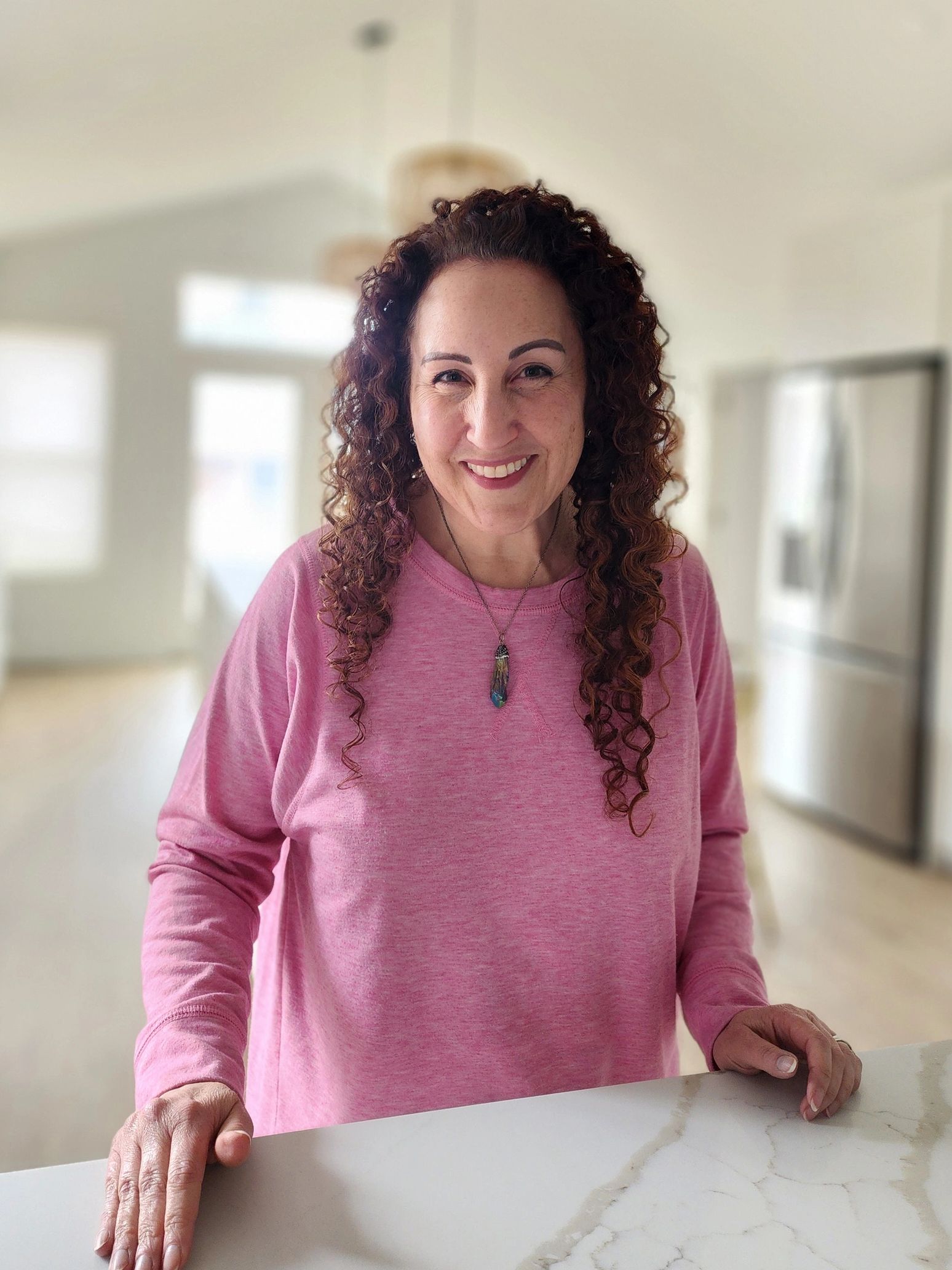 Smiling woman in a pink sweater leaning on a marble surface indoors.