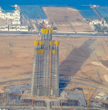 A tall skyscraper under construction casting a large shadow over a desert area.