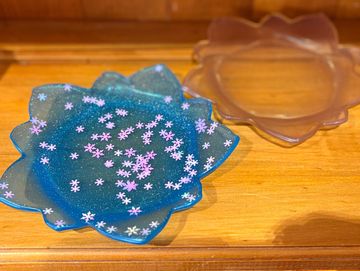 Two decorative flower-shaped trays on a wooden surface, one blue with pink speckles and one translucent.
