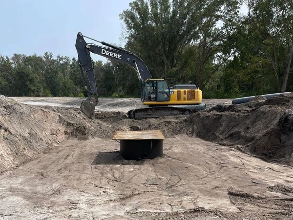 Excavator digging a large pit with pipes and trees in the background.