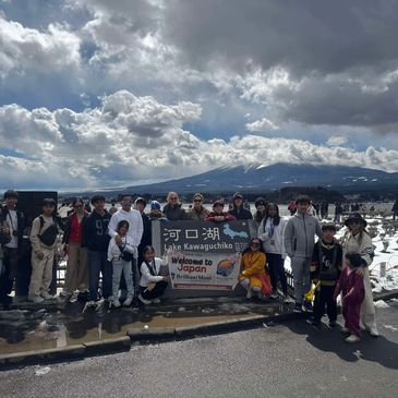 Group photo at Lake Kawaguchiko with snowy landscape and cloudy sky.