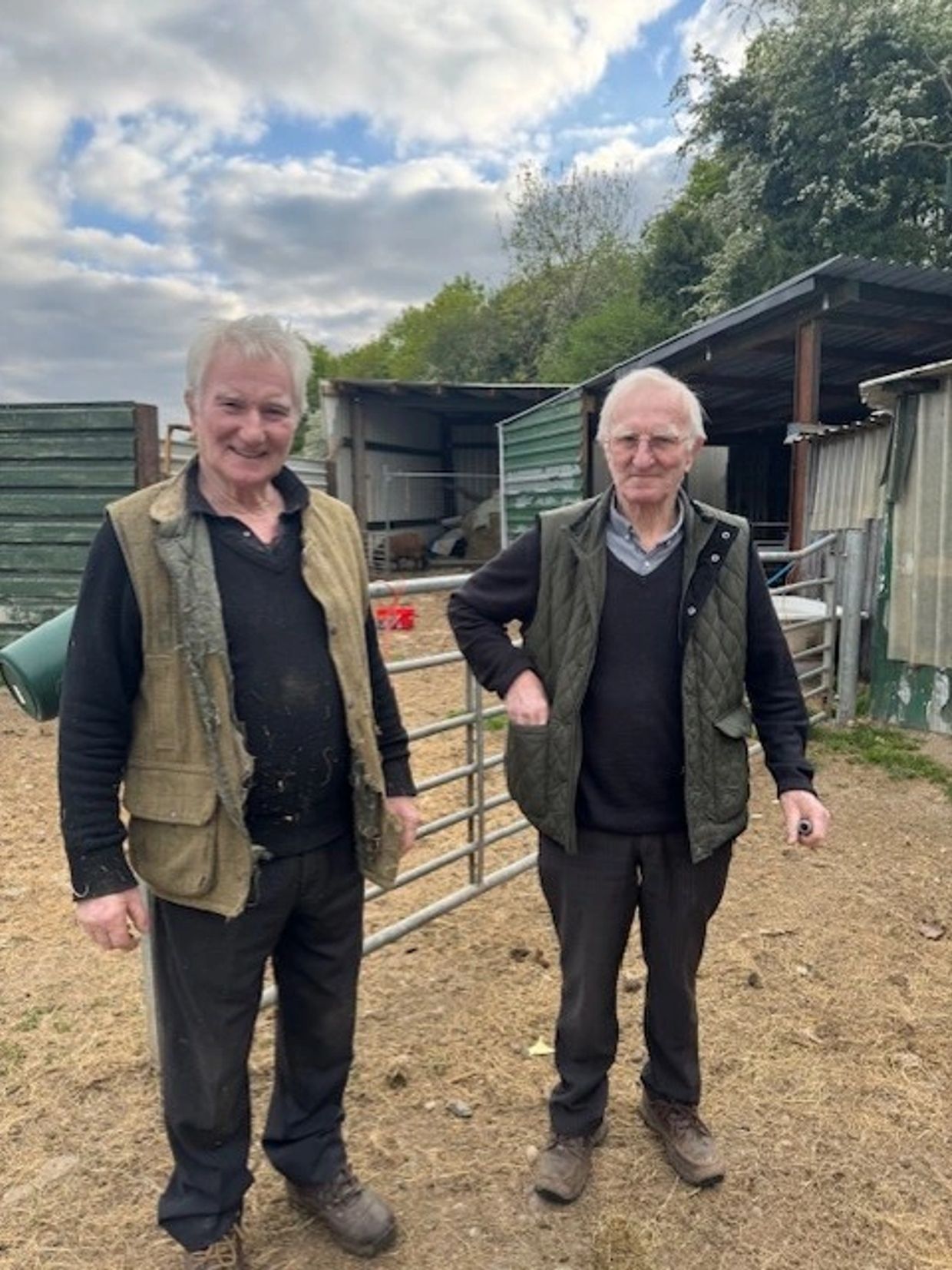 Two elderly men standing outdoors near a farm gate.