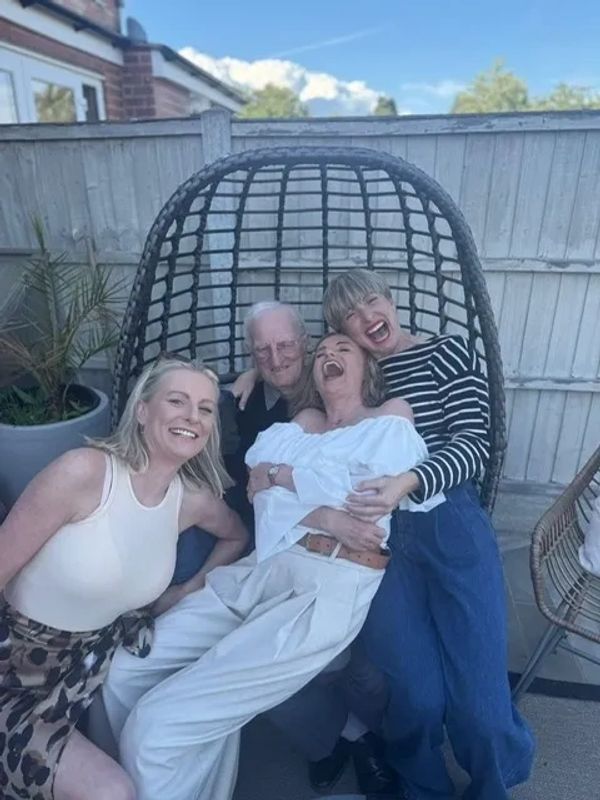Four people laughing together on a patio chair outdoors.