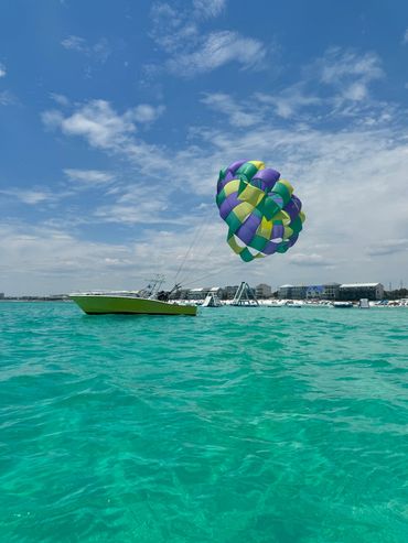 Boat towing a colorful parasail over turquoise sea near a beach resort.