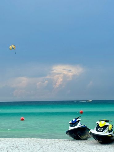 Jetskis on a sandy beach with a parasailer in the sky over turquoise water.