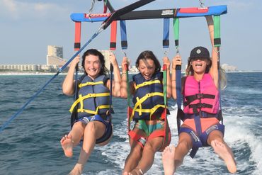 Three women smiling while parasailing over the ocean near a coastal city.